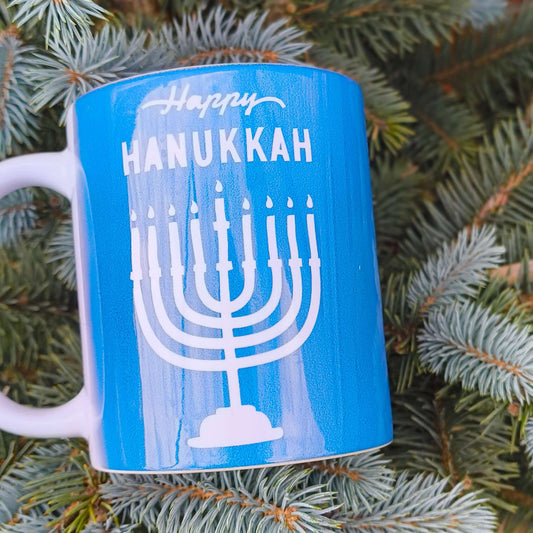 Happy Hanukkah mug featuring a menorah design on a blue background among evergreen branches.