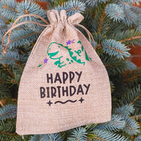 Close-up of hessian Happy Birthday gift bag with green dinosaur and purple stars, rustic design on blue spruce tree.