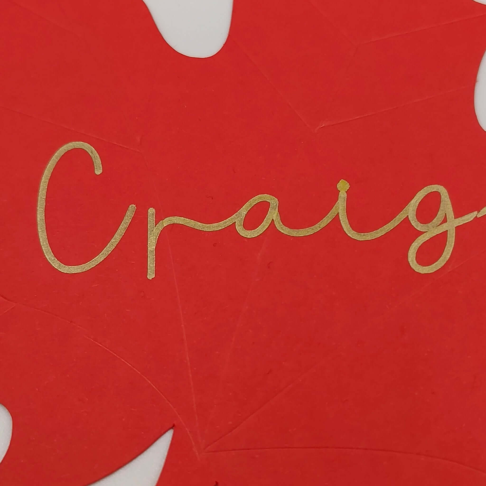 Close-up of a handmade red maple leaf place card with Craig written in shimmering gold script, showing fine detail of the cardstock texture.