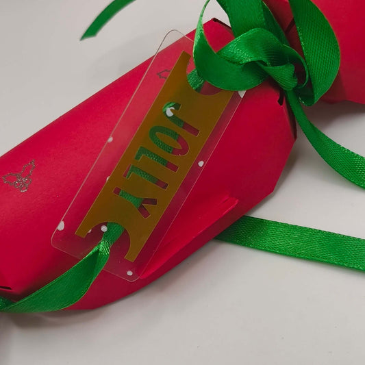 Close-up of red Christmas cracker with gold ‘JOLLY’ tag tied with green ribbon.
