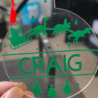 Close-up of personalised acrylic dinosaur Christmas bauble in green vinyl, featuring Santa's sleigh and festive trees.
