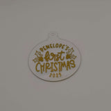 Personalised first Christmas bauble in white acrylic with gold text and holly design on plain background.