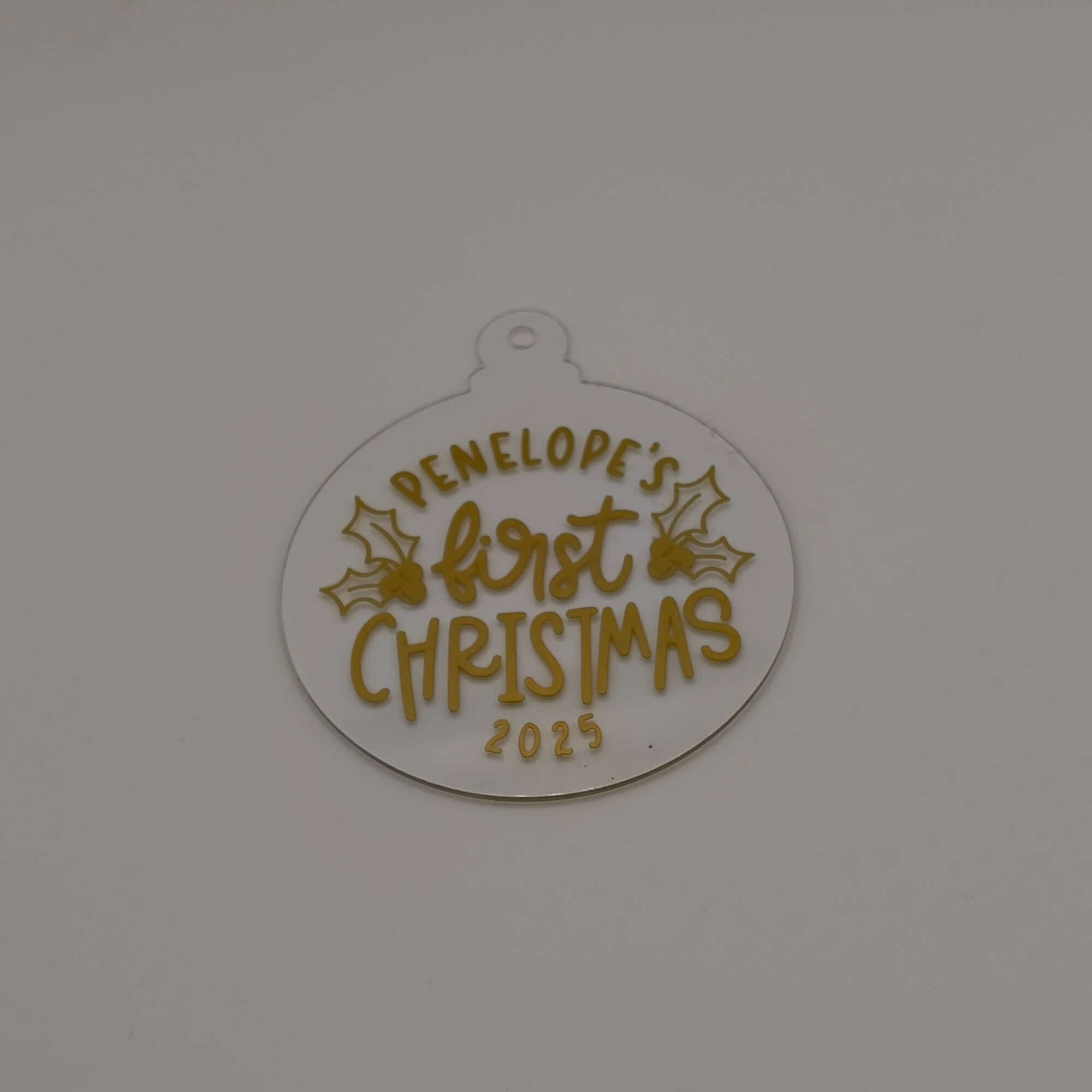 Personalised first Christmas bauble in white acrylic with gold text and holly design on plain background.