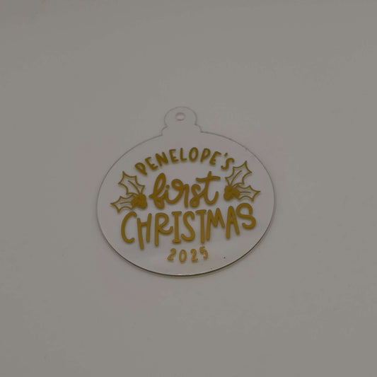 Personalised first Christmas bauble in white acrylic with gold text and holly design on plain background.