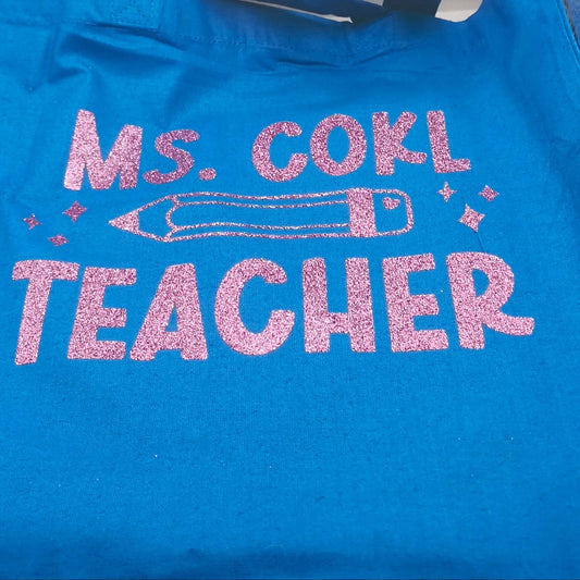 Blue tote bag with pink glitter lettering "Ms. Cohl Teacher" and pencil artwork, photographed on a neutral surface.