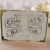Handmade white and gold baptism card with cut-out lettering reading 'Congrats on Your Baptism', displayed on a soft pink surface with a white rose in the background.