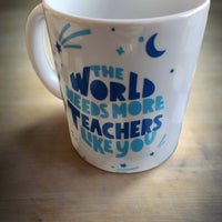 A white ceramic mug resting on a wooden surface. The mug features a motivational message in blue and teal text that reads, "THE WORLD NEEDS MORE TEACHERS LIKE YOU." Around the text are decorative elements, including stars, a crescent moon, and sparkles, adding a whimsical and inspiring touch. Perfect for celebrating and showing appreciation for teachers.