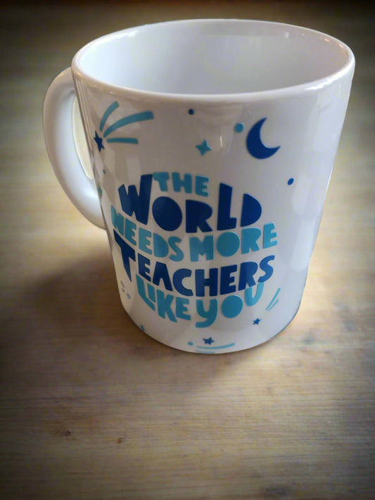 A white ceramic mug resting on a wooden surface. The mug features a motivational message in blue and teal text that reads, "THE WORLD NEEDS MORE TEACHERS LIKE YOU." Around the text are decorative elements, including stars, a crescent moon, and sparkles, adding a whimsical and inspiring touch. Perfect for celebrating and showing appreciation for teachers.
