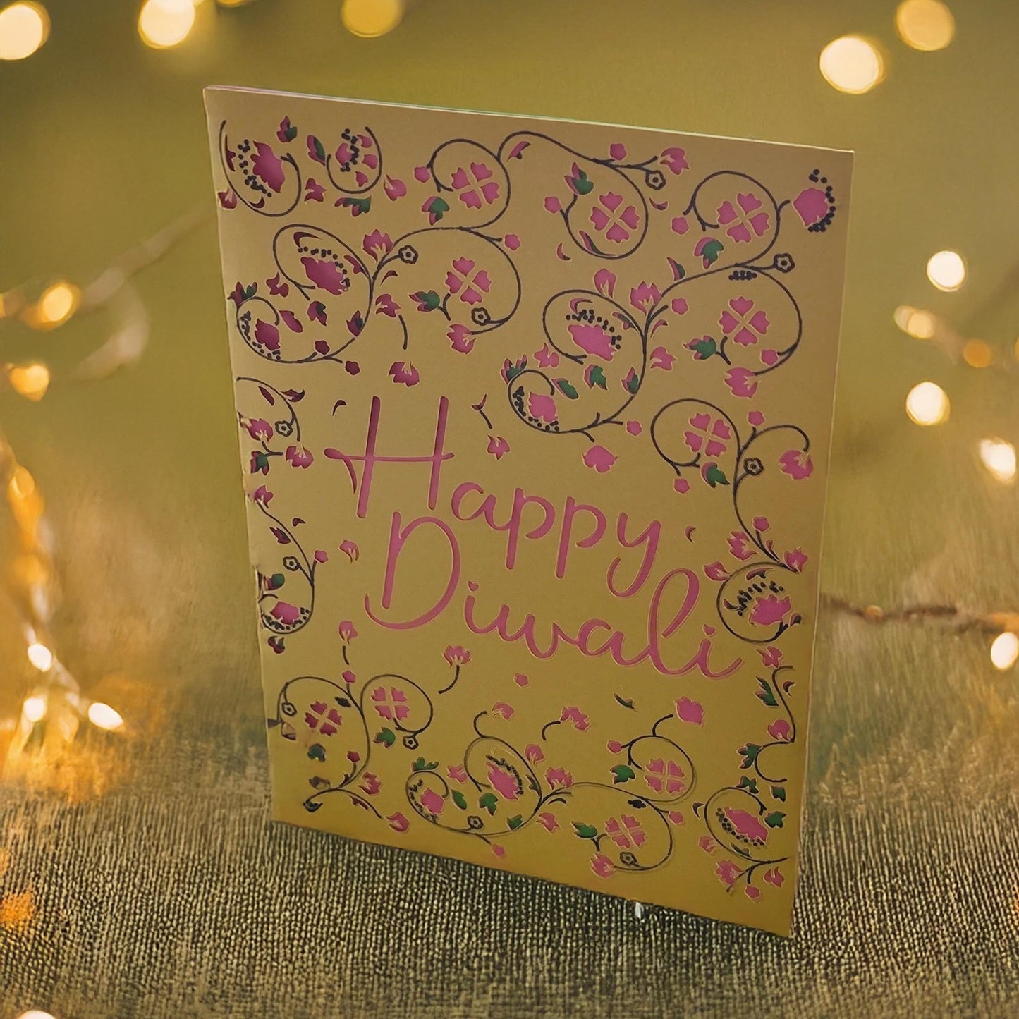 Elegant Happy Diwali card with gold background, pink cut-out text, and floral patterns, photographed with glowing diya lights.