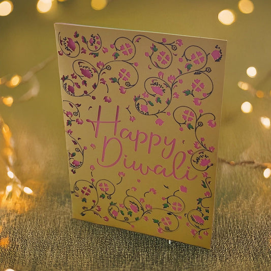 Elegant Happy Diwali card with gold background, pink cut-out text, and floral patterns, photographed with glowing diya lights.