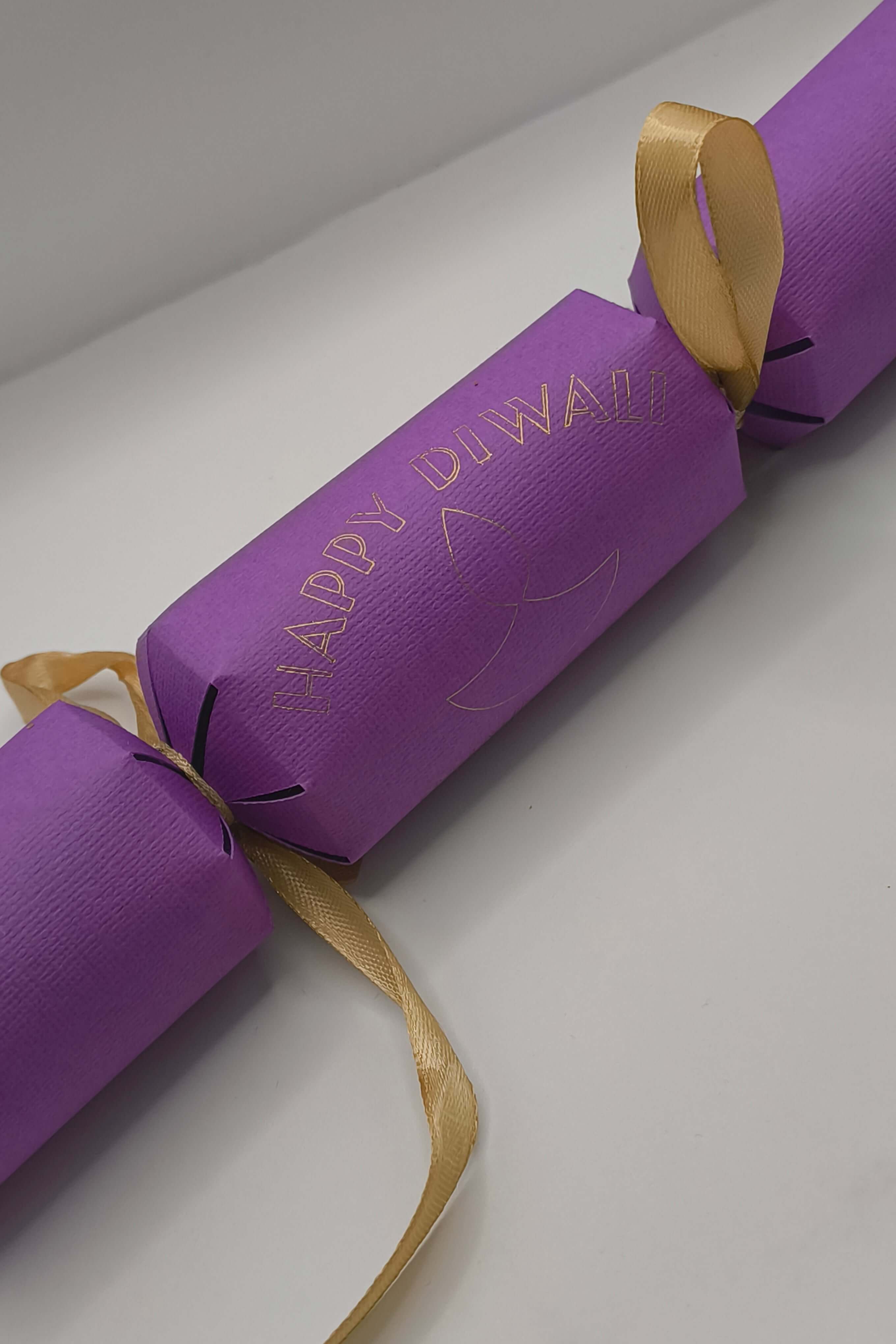 Handmade purple cracker close-up showing gold foiled “Happy Diwali” text with diya design in the centre.