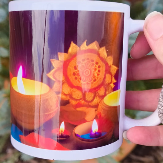 White ceramic mug featuring a vibrant Diwali design with glowing clay oil lamps (diyas) and a golden mandala pattern, held outdoors near evergreen plants.