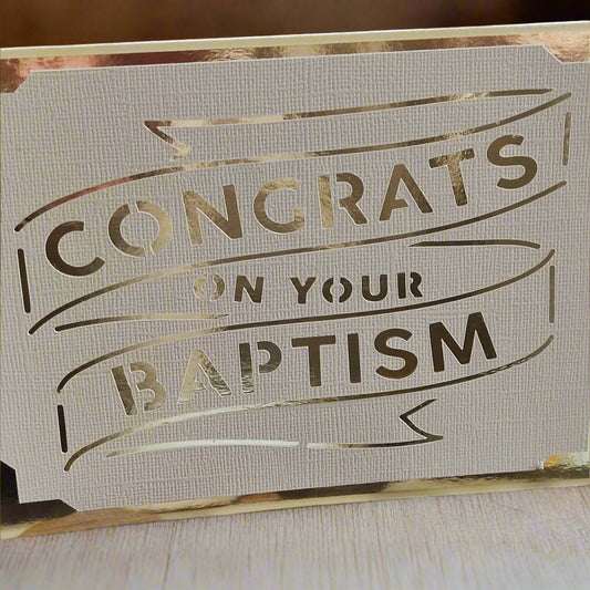 Elegant handmade baptism card with metallic gold foil text reading 'Congrats on Your Baptism', cut into textured white cardstock and displayed on a light wood surface.