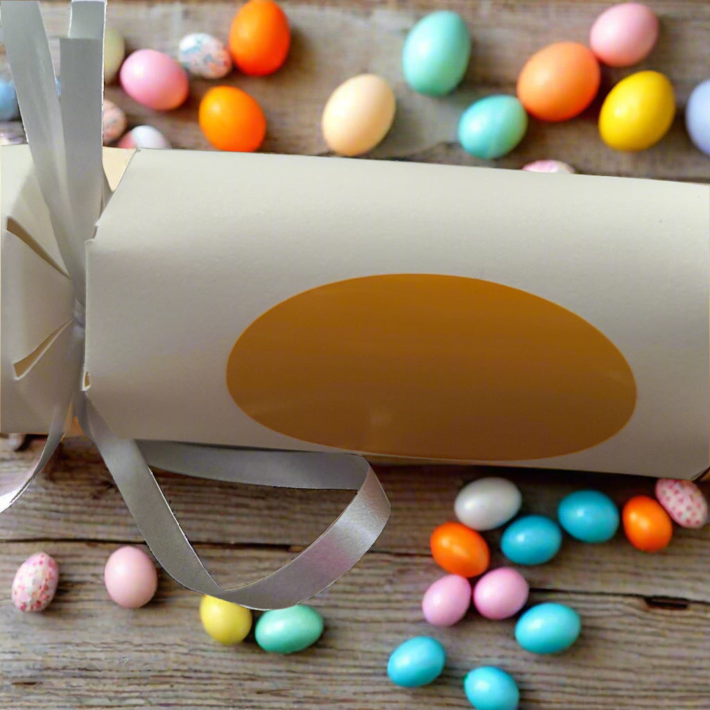 Handmade cream cardstock Easter cracker with orange oval detail, tied with silver ribbon, shown on rustic wood surrounded by colourful mini eggs.