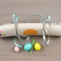 Cream Easter chick cracker with orange oval belly and silver ribbon ties, displayed flat on a table beside pastel mini eggs for size reference.