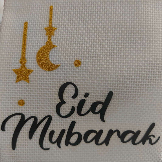 Close-up of a white textured hessian fabric with gold glitter moon and star decorations and the black text 'Eid Mubarak' in elegant script.