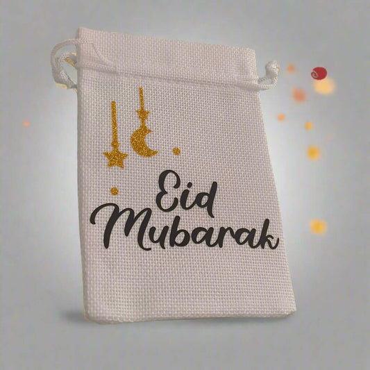 White hessian drawstring bag with 'Eid Mubarak' text in black and gold glitter crescent moon and stars, placed against a soft grey background with glowing orange bokeh lights.