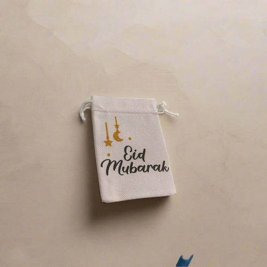White hessian drawstring bag with 'Eid Mubarak' in black text and gold glitter moon and star decorations, placed on a soft beige marble background with subtle shadows.