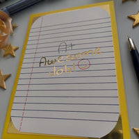 A piece of lined paper with a yellow border displaying the handwritten note “A+ Awesome Job!” in black and gold ink, accompanied by a red-circled smiley face. A pen sits to the right, with gold star embellishments adding a celebratory touch.