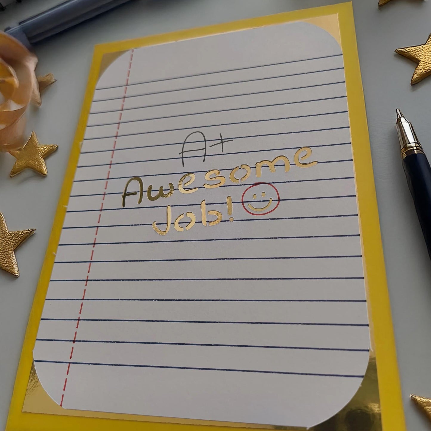 A piece of lined paper with a yellow border displaying the handwritten note “A+ Awesome Job!” in black and gold ink, accompanied by a red-circled smiley face. A pen sits to the right, with gold star embellishments adding a celebratory touch.