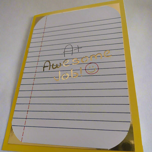 Encouragement greeting card with A+ Awesome Job! message in gold foil on lined paper, mounted on yellow card, photographed from a low side angle.