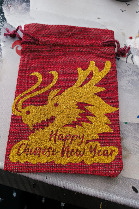 Festive red hessian drawstring bag with gold glitter dragon print and New Year message, flat-lay style.
