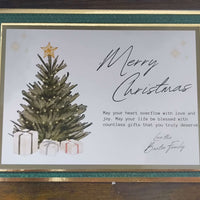 Flatlay of a luxury handmade Christmas card featuring a green textured border with gold foiling. The centre design shows a lit Christmas tree topped with a gold star and surrounded by wrapped gifts. The card includes the scripted message “Merry Christmas” and a printed sentiment: “May your heart overflow with love and joy. May your life be blessed with countless gifts that you truly deserve.” Signed “Love the Baxter Family.”