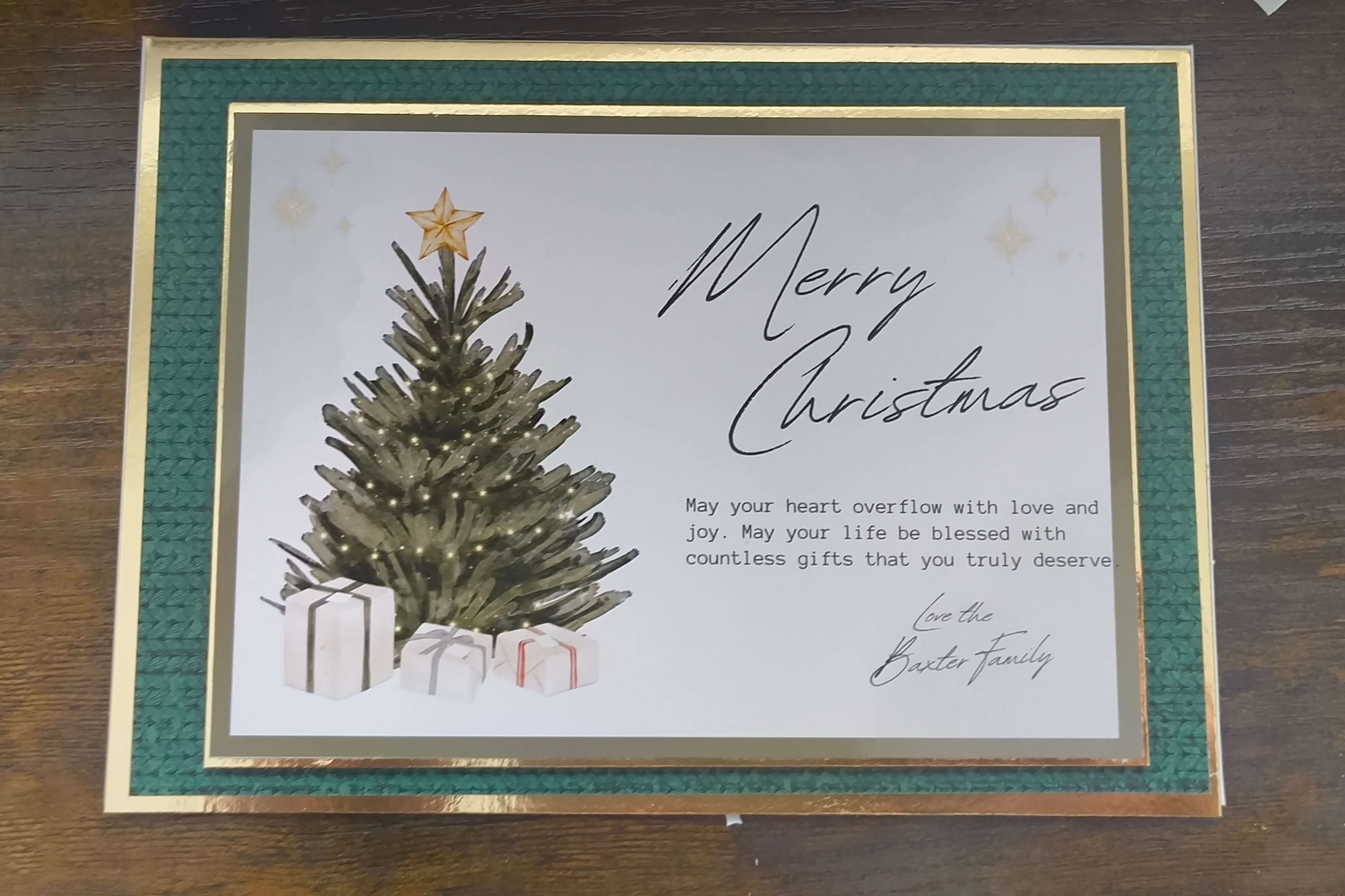 Flatlay of a luxury handmade Christmas card featuring a green textured border with gold foiling. The centre design shows a lit Christmas tree topped with a gold star and surrounded by wrapped gifts. The card includes the scripted message “Merry Christmas” and a printed sentiment: “May your heart overflow with love and joy. May your life be blessed with countless gifts that you truly deserve.” Signed “Love the Baxter Family.”