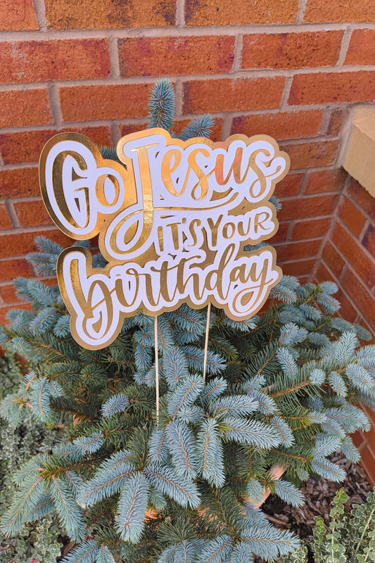 Gold and white “Go Jesus It’s Your Birthday” cake topper displayed on a small blue spruce tree against a brick wall.
