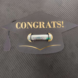 Black graduation cap-shaped money holder with “CONGRATS!” in gold text, holding a rolled banknote, placed on a textured dark background.