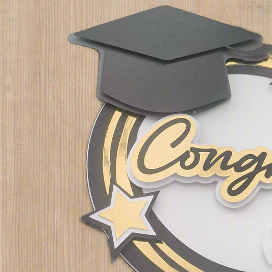 Handmade graduation card on a woodgrain background, featuring a black mortarboard, gold foiled “Congrats” text, and layered gold star embellishments. Ideal for graduation crafts, scrapbook pages, and handmade greeting cards.