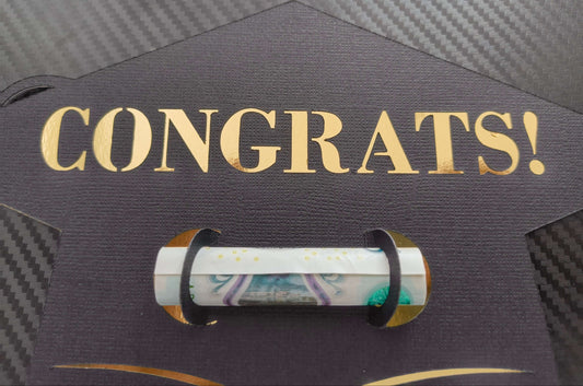 Close-up of black graduation cap-shaped money holder with gold “CONGRATS!” lettering and rolled banknote held securely in the centre slot.