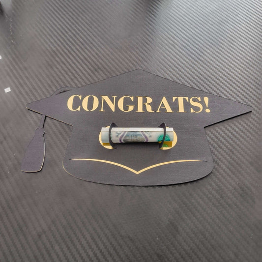 Black graduation cap-shaped money holder with gold “CONGRATS!” text, holding a rolled banknote in the centre, displayed on a dark textured background.