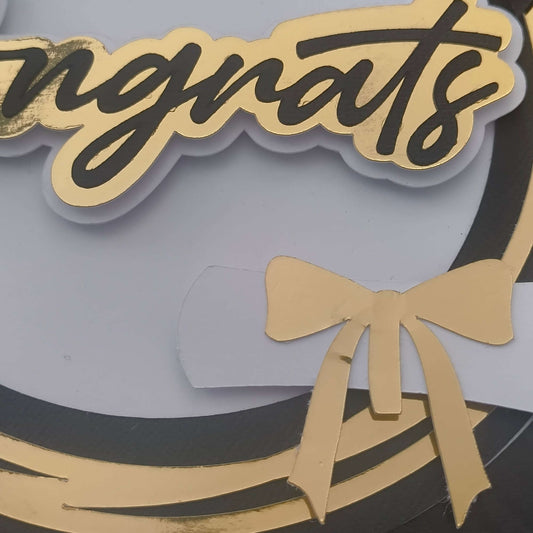 Close-up detail of gold foiled “Congrats” lettering and rolled diploma with golden bow on a handmade graduation card topper. Features elegant layered cardstock in black, white, and gold for a premium finish.