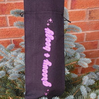 Upright black drawstring wine bag with glitter pink “Groovy & Boozy” lettering, positioned on a pine bush by brick wall.