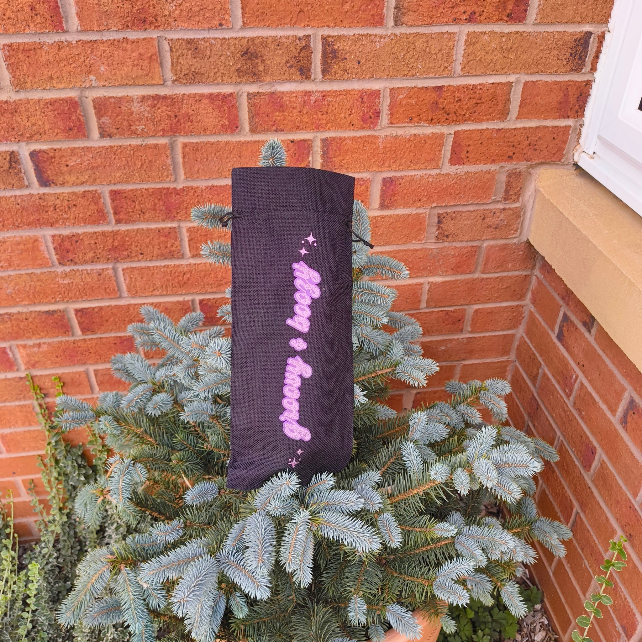 Black drawstring wine bag with glitter “Groovy & Boozy” design displayed on top of a small pine bush against brick wall.