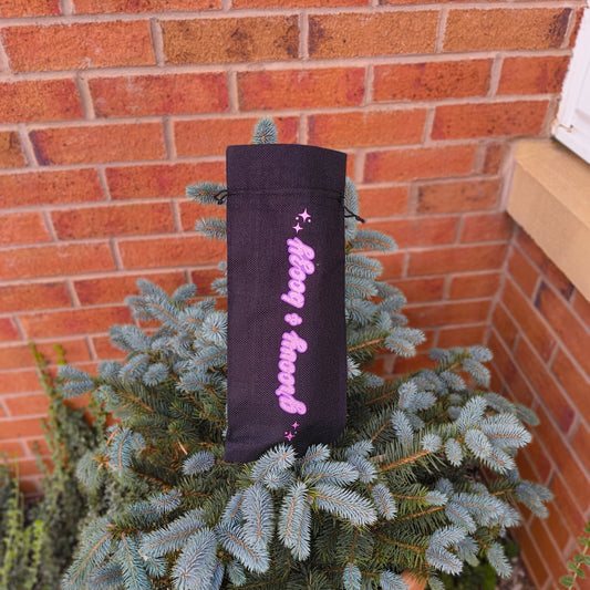 Glittery pink “Groovy & Boozy” wine bag displayed outdoors on a pine tree, sparkly lettering standing out on black fabric.
