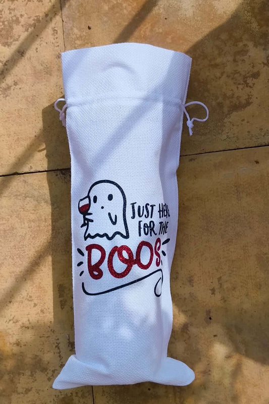 Flat-lay of a Halloween wine bag featuring ghost illustration and glitter red "BOOS" lettering, photographed outdoors on stone tiles.