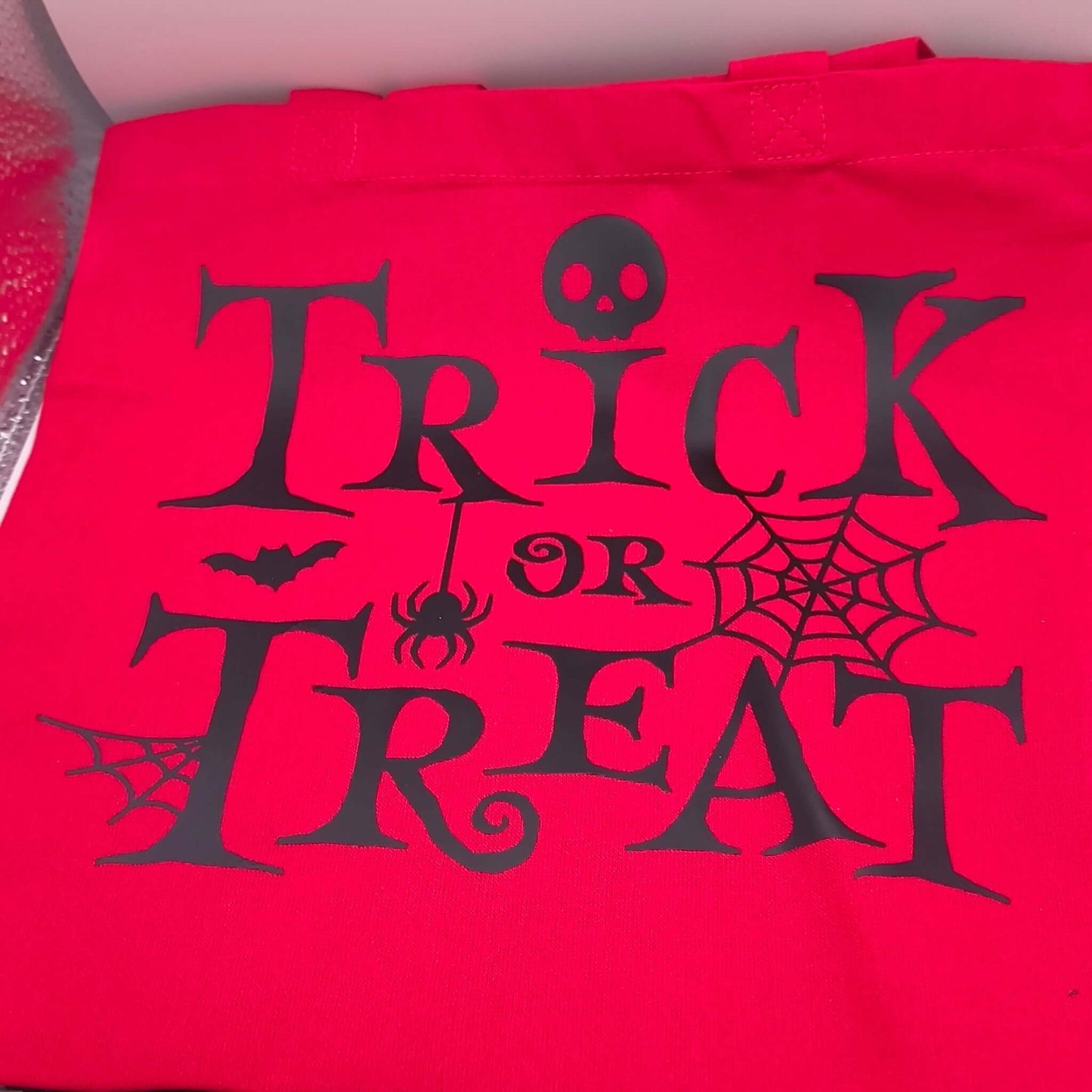 Red Halloween trick or treat tote bag with black spooky font, skull, spider, bat and cobweb design, perfect for kids to collect sweets.