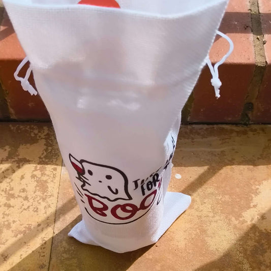 White drawstring wine gift bag with a ghost and "Just Here for the Boos" design, standing upright on outdoor tiles.
