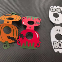 Flat-lay close-up of three Halloween-themed lollipop holders in pumpkin orange, red cauldron with green drip, and white ghost with "BOO" text, arranged side by side on a black carbon-textured background.