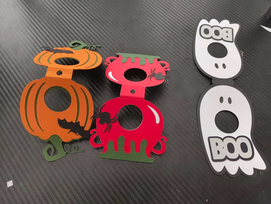 Flat-lay close-up of three Halloween-themed lollipop holders in pumpkin orange, red cauldron with green drip, and white ghost with "BOO" text, arranged side by side on a black carbon-textured background.
