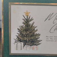 Close-up of the left side of a handmade Christmas card featuring a decorated tree with warm white lights and a gold star topper. The base of the tree is surrounded by wrapped presents in white paper with red and black ribbons. The card is bordered in deep green textured cardstock and gold foil trim.