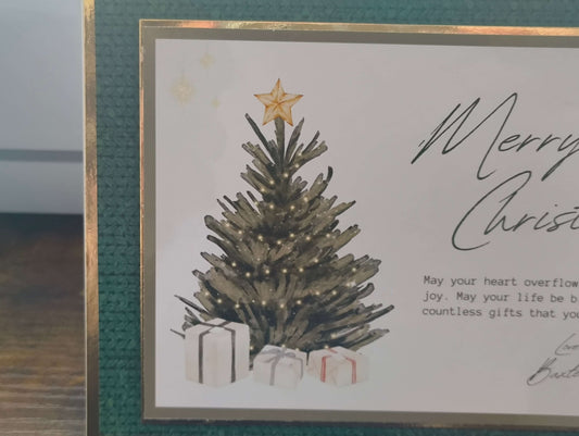Close-up of the left side of a handmade Christmas card featuring a decorated tree with warm white lights and a gold star topper. The base of the tree is surrounded by wrapped presents in white paper with red and black ribbons. The card is bordered in deep green textured cardstock and gold foil trim.