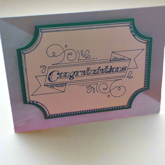 A textured ivory congratulations card with a teal frame and ornate banner lettering. Angled shot shows layered cardstock detail and standard fold size.