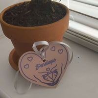 Personalised heart-shaped baby gift tag with name 'Penelope' and ribbon attached, resting near a potted plant.
