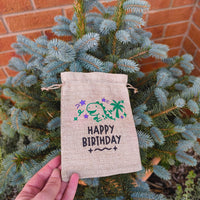 Handmade Happy Birthday hessian bag with dinosaur design, held by hand in front of blue spruce tree and terracotta pot.