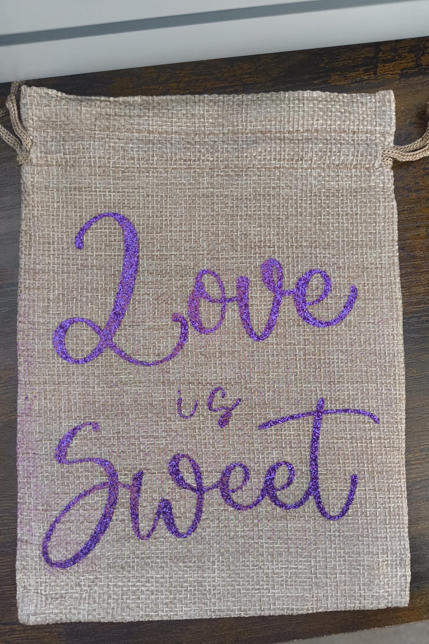 Handmade hessian bag with a natural woven texture and bold purple glitter script reading “Love is Sweet.” Sturdy and reusable with a simple drawstring fastening.