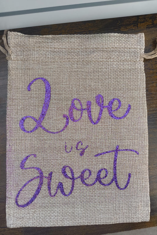 Handmade hessian bag with a natural woven texture and bold purple glitter script reading “Love is Sweet.” Sturdy and reusable with a simple drawstring fastening.