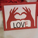 Handmade love card in red and white with heart hands design and the word LOVE on the front, displayed against a background of red hearts.